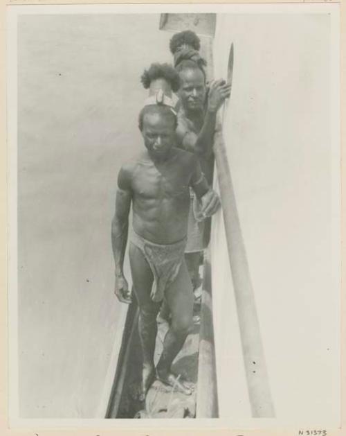 Two men wearing headdresses, standing in canoe