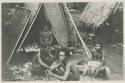 Woman and three men sitting in front of thatched structure