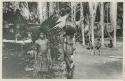 Two men standing in front of trees, children in background