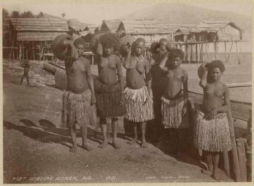 Group of women standing on beach, with pile dwellings in background