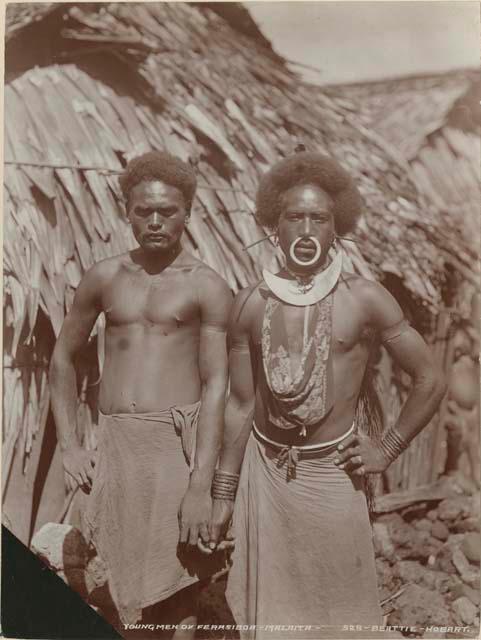 Two men standing in front of a thatched structure, one with a nose ring