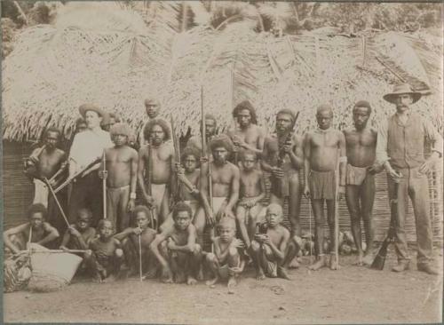 Group of men and youth holding rifles and sticks by thatched structure