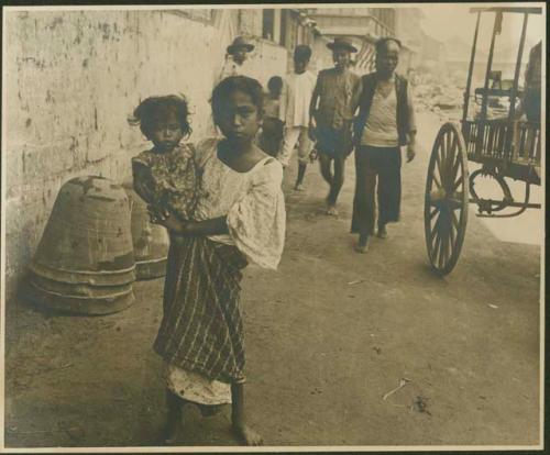 Girl holds baby on hip, men and wagon behind