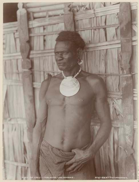 Man with shell necklace standing in front of thatched structure