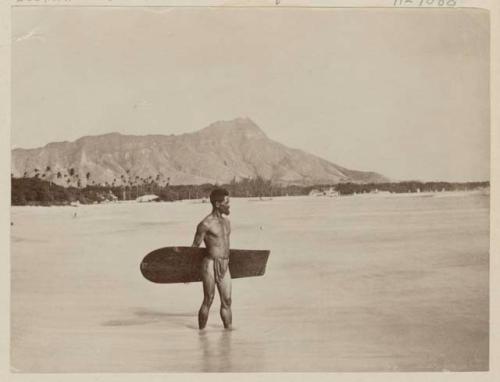 Man holding swimming board in shallows, shoreline and mountains in background