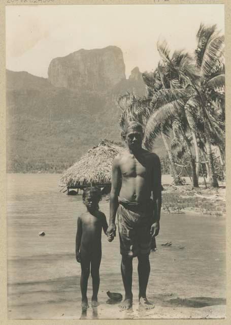 Man and child standing on shore, with thatched structure in background