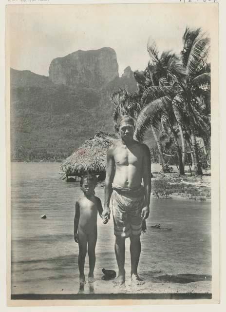 Man and child standing on shore, with thatched structure in background