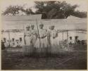 Women in ceremonial dress standing in front of long shelter