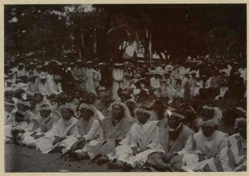 Line of women sitting, with large crowd in background