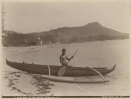 Man on shoreline in outrigger canoe, Diamond Head in background