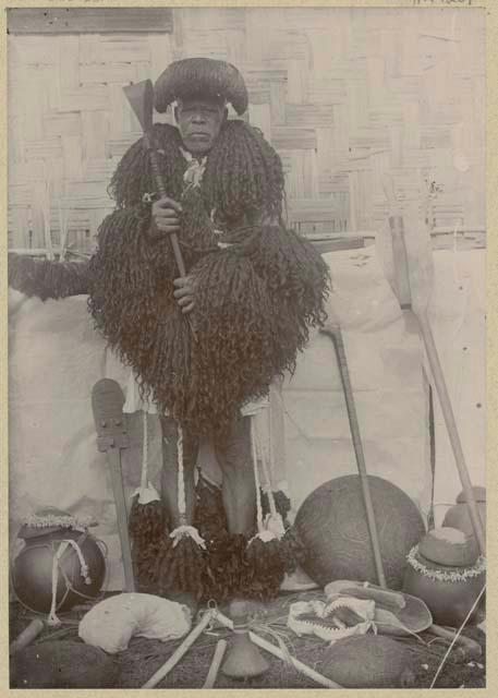 Man standing among objects dressed in a human hair cape and apron