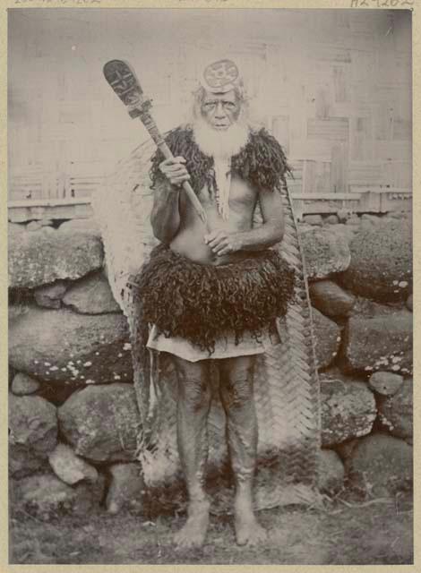 Man with a club dressed in human hair skirt and shoulder cape