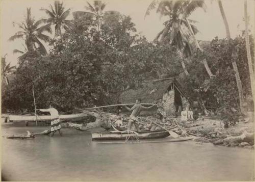 Man in canoe throwing spear, people on shore outside building