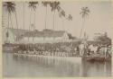 Men in long boat next to shoreline with people, fence, and building