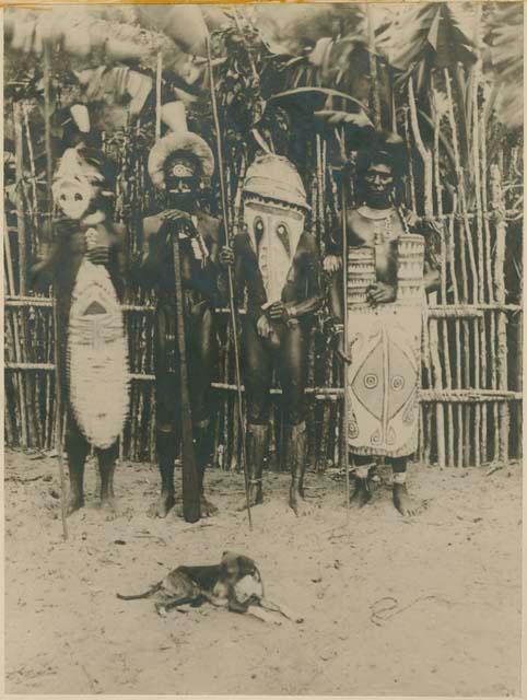 Men standing with body shield carvings, a dog lies at rest in foreground