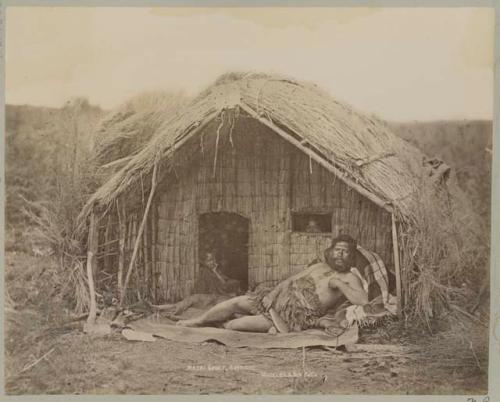 Man lounging in front of straw hut, people in door way and windows