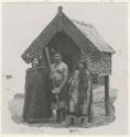 Three people outside small house carved with stone implements