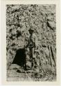 Man standing in front of communal house with rifle in his right hand.