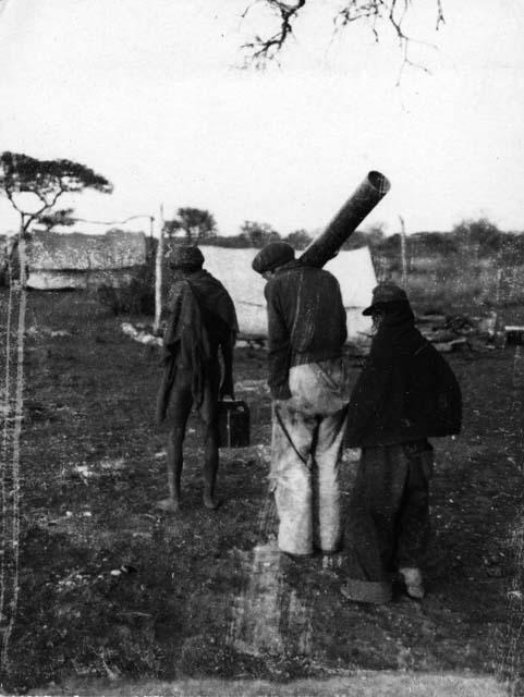 Three people, including two wearing Western clothes, walking, carrying equipment, with tents in the background