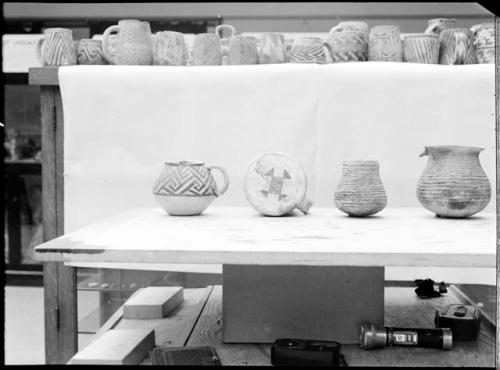 Four pottery vessels, smaller pots & mugs above on shelf.