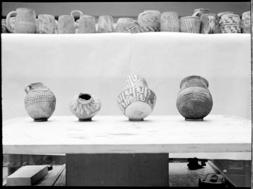 Four pottery vessels, smaller pots & mugs above on shelf.