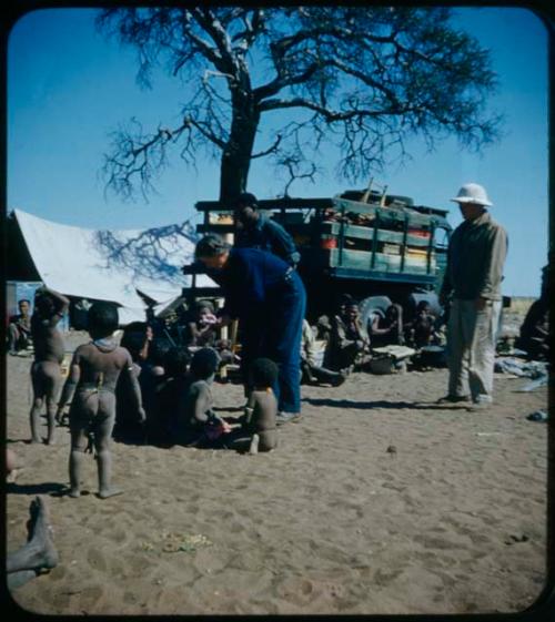 Expedition: Lorna Marshall,  Laurence Marshall, and Kernel Ledimo handing out gifts to people, with a tent and a truck in the background