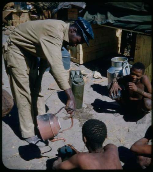 Expedition: Expedition member bending over to pour water from a kettle into an ostrich eggshell held by a seated man, with other people watching