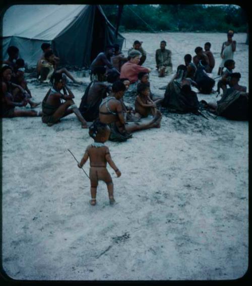 Expedition: Boy standing, with a group of people, including Lorna Marshall, sitting by a tent in the background