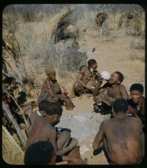 Groups: Group of people sitting in front of a skerm, including a woman using a mortar and a man drinking from an ostrich eggshell