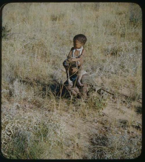 Food Gathering, Children: Woman squatting to gather something, carrying a child on her shoulders