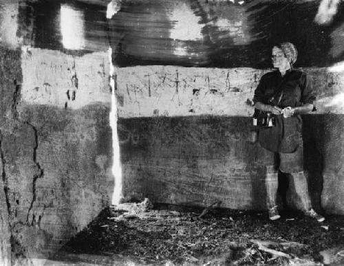 Elizabeth Marshall Thomas standing next to drawings made by children on a wall inside a hut