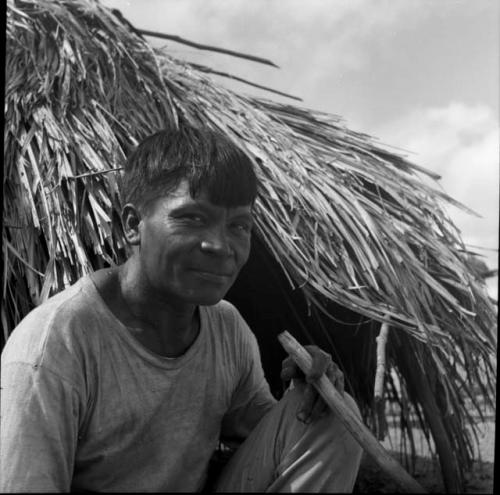 Portraits of people: man in front of thatched building holding stick