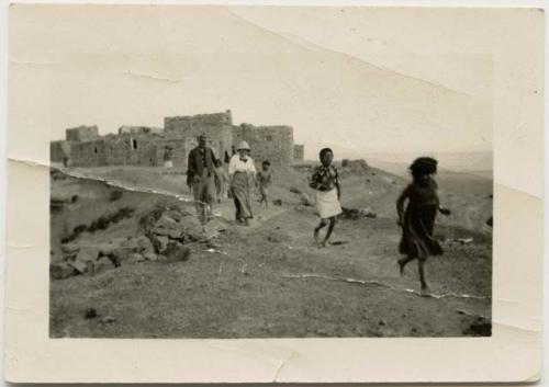 Hopi people walking with Grace Nicholson, with pueblo in background