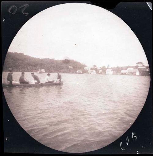 Canoe with people on water, buildings on shore behind