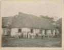 Crowd of men and local dignitaries lined up in front of thatched-roof buildings