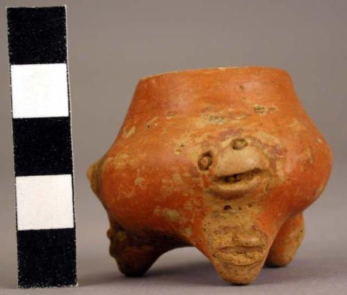 Miniature red tripod vessel with a plastic head above each leg and motif on leg