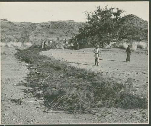 Men standing next to row of brush laid out on sand (print is a cropped image)