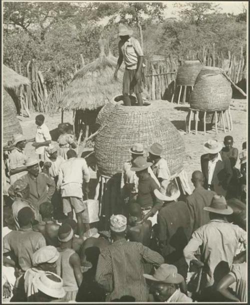 Group of people standing in front of storage baskets, and a man standing on top of one (print is a cropped image)