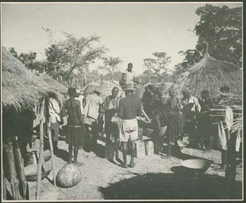 Group of people standing in front of storage basket, and man sitting on top of one holding a can (print is a cropped image)