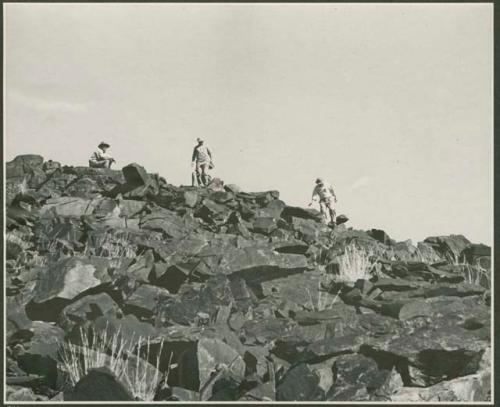 Four men standing and sitting at the top of a rocky hill (print is a cropped image)