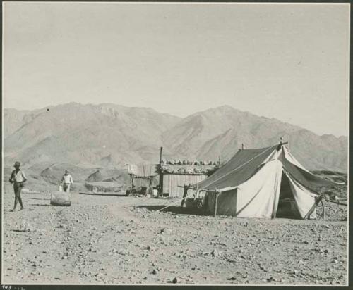 Men walking through a camp with a tent and corrugated metal shed (print is a cropped image)