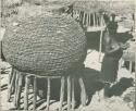 Woman standing next to a large basket on a stand (print is a cropped image)