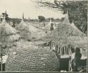 People standing next to large baskets and thatched roofs (print is a cropped image)