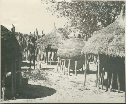 Woman standing next to storage baskets inside kraal (print is a cropped image)