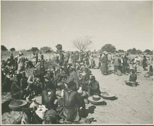 Group of people sitting and standing, with baskets of grain on the ground near them (print is a cropped image)