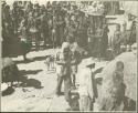 People standing next to huts, with government officials in background (print is a cropped image)