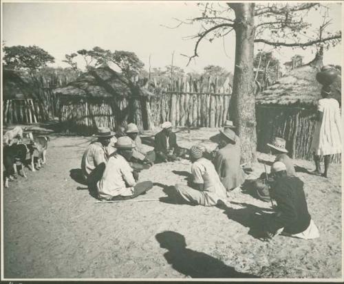 Men sitting near a tree, and a woman carrying a pot on her head (print is a cropped image)