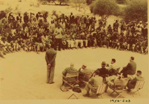 Indaba at Omajetti, Herero chief, with a large group of people sitting and standing behind him, addressing expedition members, including Colonel Hoogenhout, John Neser, Laurence Marshall and John Marshall