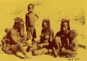 Group of women making a necklace, with children sitting and standing with them