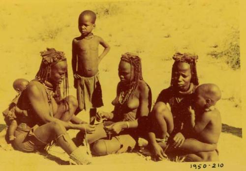 Group of women making a necklace, with children sitting and standing with them